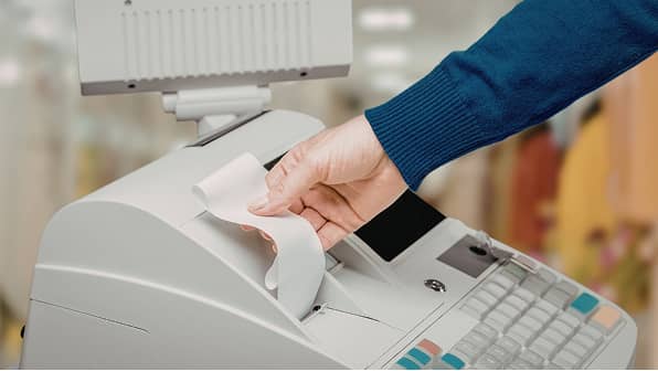 店舗のレジカウンターで、店員がレジスターから発行されたレシートを手で取り出す瞬間のクローズアップ。青い袖の服を着た店員の手元と、白いレジスターのキーボードやモニターが映し出されている。