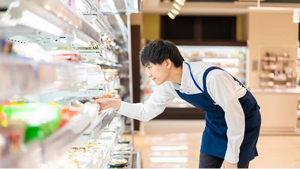 スーパーマーケットの冷蔵棚で、商品を丁寧に陳列している若い男性店員。白いシャツに紺色のエプロンを着用し、真剣な表情で棚の奥まで商品を確認しながら並べている。背景には明るい店内の様子が見える。