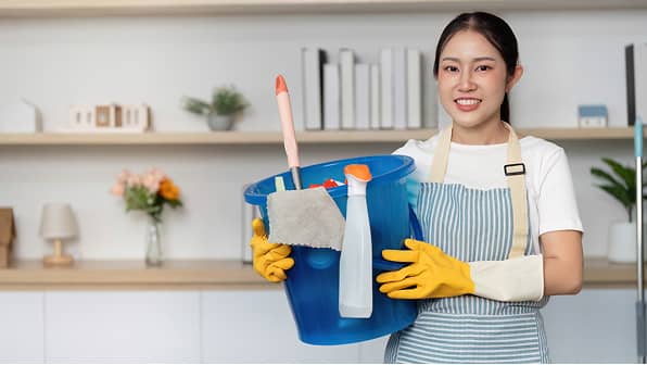 清掃用具が入った青いバケツを抱え、笑顔でカメラを見る女性スタッフ。ストライプのエプロンと黄色いゴム手袋を着用し、バケツにはスプレーボトルやブラシが入っている。背景には整理された室内の棚が見える。