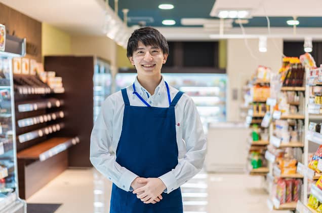 明るい店内で、青いエプロンと白いシャツを着用した男性店員が、カメラに向かって爽やかに微笑んでいるポートレート。清潔感のある接客イメージを伝えます。