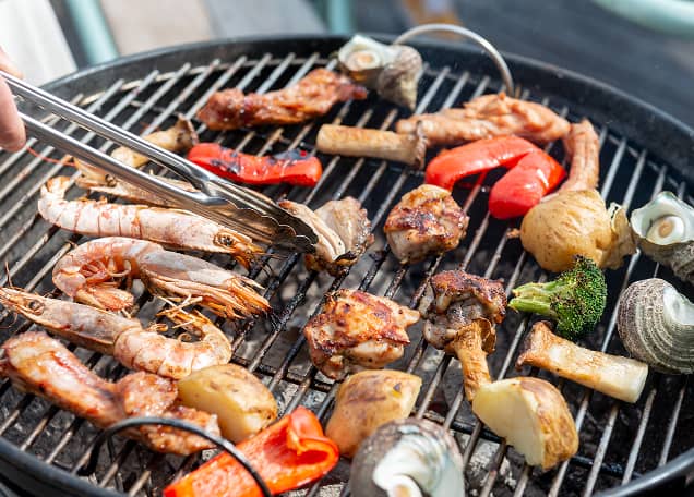 網の上で豪華な食材が焼かれているバーベキューの様子。大きなエビ、厚切りの肉、サザエ、彩り豊かなパプリカやブロッコリーが並び、トングで食材を裏返している食欲をそそるシーン。