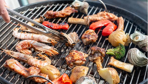 豪華な食材が並ぶバーベキューの調理風景。海鮮、肉、彩り豊かな野菜が炭火でじっくりと焼き上げられており、野外での食事の楽しさと美味しさを強調しています。