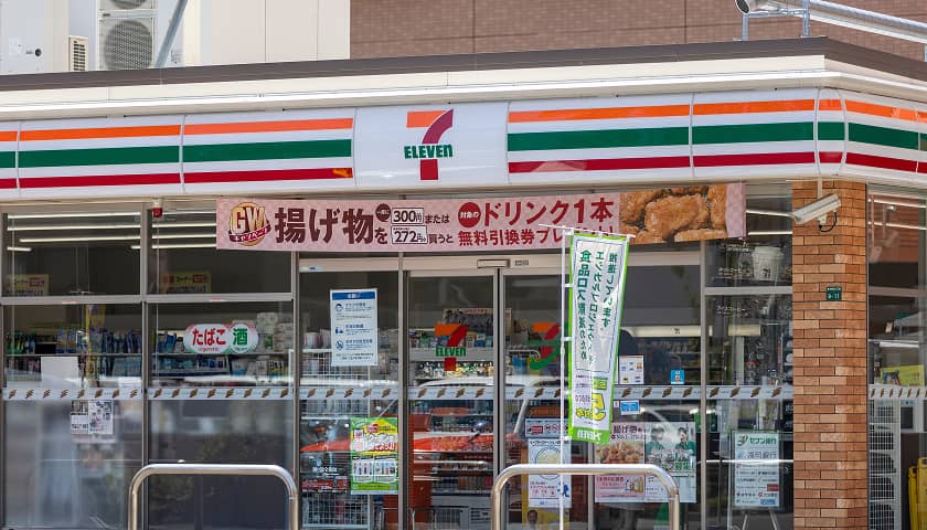 昼間のセブン-イレブン店舗入口付近の外観写真。キャンペーン告知の幕やのぼり旗が設置されており、賑やかで利便性の高い店舗の様子を伝えています。