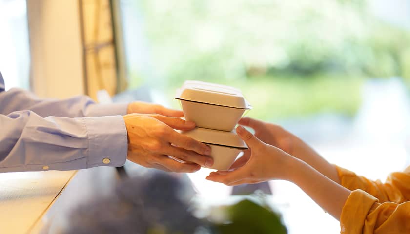テイクアウト商品の受け渡しシーン。スタッフからお客様へ、温かい料理が入った容器を直接手渡す様子が、サービスの丁寧さと手軽さを強調しています。